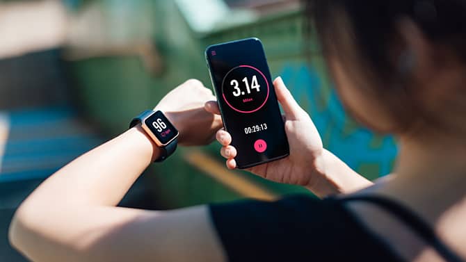 Eine Frau, die eine Fitness-App auf ihrem Smartphone und ihrer Smartwatch nutzt. Fitness-Tracking-Apps sind ein Beispiel für die positiven Auswirkungen moderner Technologien auf die Gesundheit.