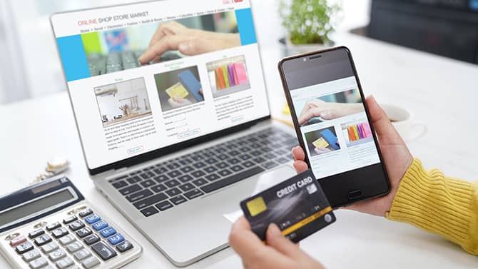Nahezu jede Online-Aktivität kann zu Ihrem digitalen Fußabdruck beitragen. Das Bild zeigt eine Person mit einem Telefon und einer Kreditkarte in der Hand, die vor einem Computerbildschirm mit einem geöffneten Online-Shop sitzt.